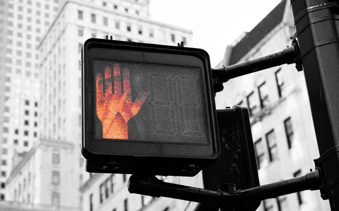 A crosswalk sign in a city. A red hand on the sign is lit up, signaling people to stop